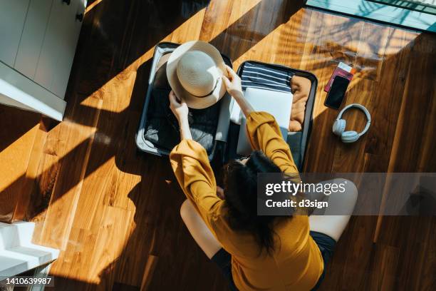 overhead view of young asian woman sitting on the floor in her bedroom, packing a suitcase for a trip. getting ready for a vacation. traveller's accessories. travel and vacation concept - viajes fotografías e imágenes de stock