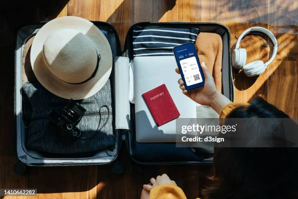 overhead view of asian woman holding smartphone showing electronic flight ticket above an open suitcase with clothings, sun hat, camera, headphones, laptop and passport on wooden floor against sunlight. traveller's accessories. travel and vacation concept - apple pay mobile payment stock-fotos und bilder