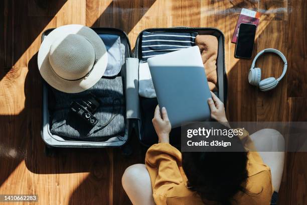 overhead view of young asian woman packing a suitcase for a trip at home. she is putting a laptop in her suitcase, with sun hat, camera, clothings, headphones, smartphone and passport by the side. getting ready for a vacation. travel and vacation concept - nueva-normalidad-concepto fotografías e imágenes de stock