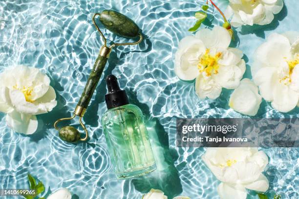 glass bottle of natural cosmetic serum with a pipette, massage essential oil and jade face roller. with white rose flowers on a background of blue water with waves. cosmetic products for body and face care. - white rose flower spa stock-fotos und bilder