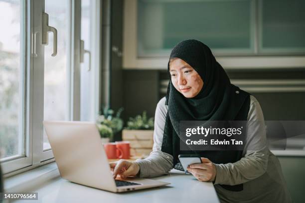 asian malay woman in hijab with vitiligo skin condition at kitchen using laptop - human skin 個照片及圖片檔