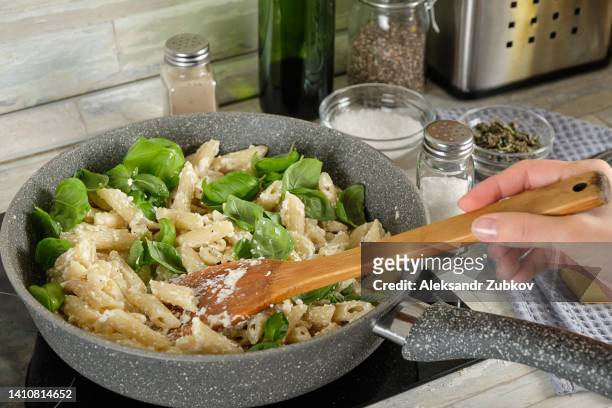 we cook italian pasta with cream sauce and basil at home. a girl or a woman cooks and stirs penne pasta in a saucepan or in a frying pan, adds herbs to it. vegetarian food. ethical consumption. step-by-step instructions, do it yourself. step 4. - one pot pasta stock-fotos und bilder