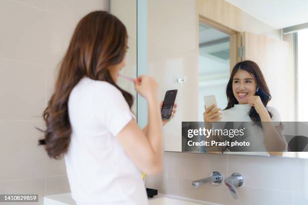 asian woman in pajamas checks e-mail on her smartphone while brushing her teeth in the bathroom to get ready for work in the morning. - brush teeth phone stock pictures, royalty-free photos & images