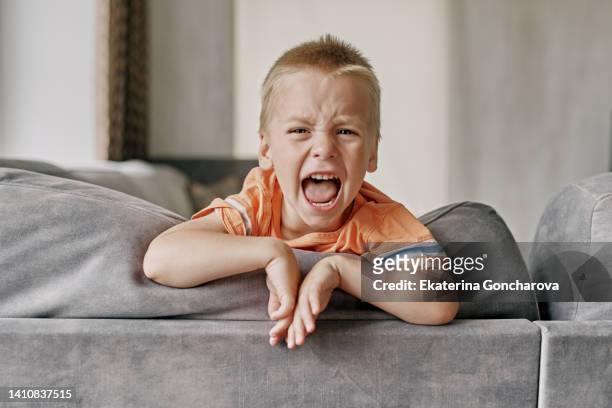 a little boy with blond hair, three years old, is sitting on the sofa and crying hysterically after kindergarten at home. parenting. - hysteria stock-fotos und bilder