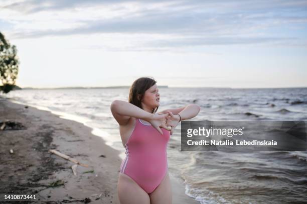 teenage girl of 12 years old with overweight in pink swimsuit does sports exercises on shore of big lake. - 12 years old girls ストックフォトと画像