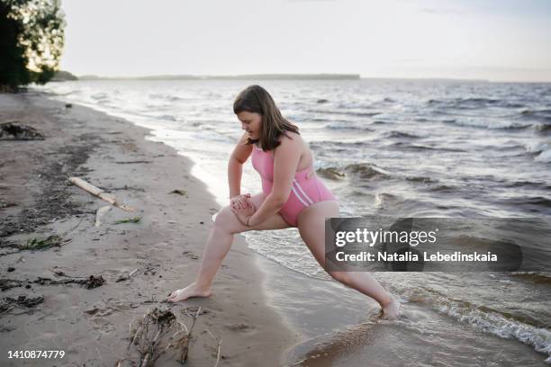 teenage girl of 12 years old with overweight in pink swimsuit does sports exercises on shore of big lake. - 12 years old girls stock-fotos und bilder