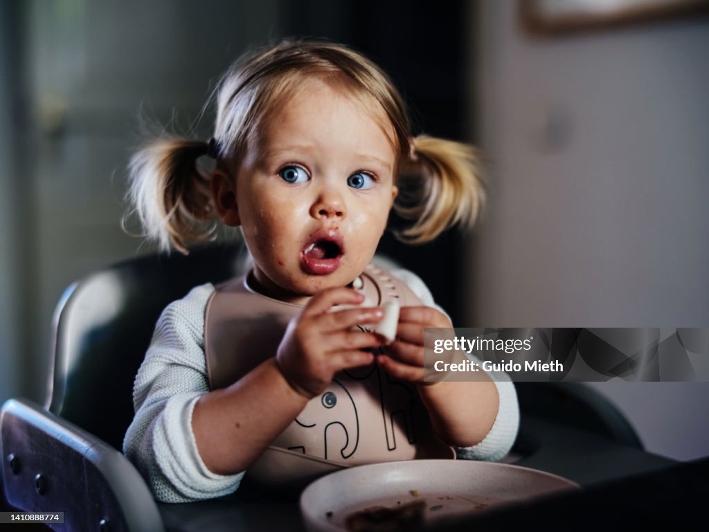 Amazed cute toddler girl with pigtails eating at a table at home.