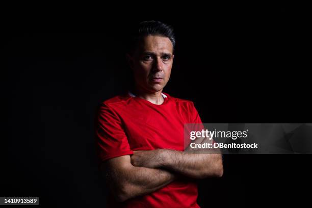 portrait of a male soccer player, wearing a red jersey, with his arms crossed, on a black background with hard shadows. concept of penumbra, world cup, professional athlete and competition. - professional sportsperson ストックフォトと画像
