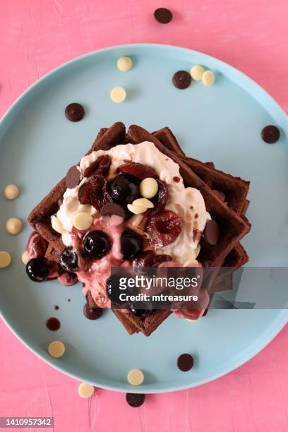 close-up image of stack of homemade chocolate waffle squares topped with melting raspberry ice cream, blueberries and cherries, milk and white chocolate chips, light blue plate, pink background, elevated view - temptation stock pictures, royalty-free photos & images