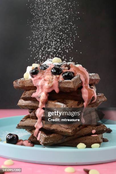 image of icing sugar sifted onto stack of homemade chocolate waffle squares topped with melting raspberry ice cream, blueberries and cherries, milk and white chocolate chips, light blue plate, pink surface, black background, focus on foreground - waffles stock pictures, royalty-free photos & images