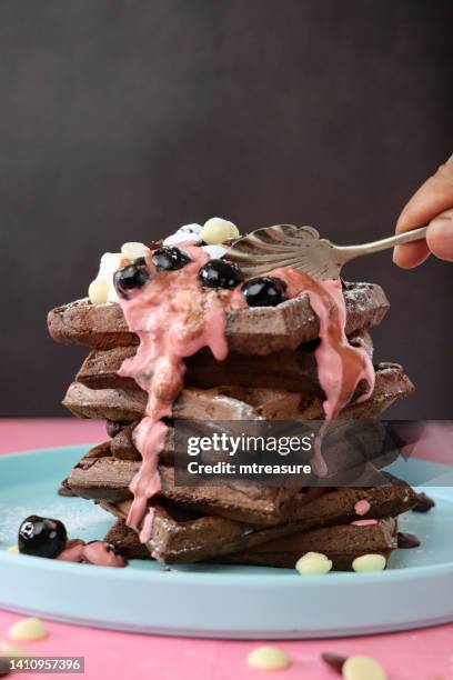 image of unrecognisable person using spoon to cut into stack of homemade chocolate waffle squares topped with melting raspberry ice cream, blueberries and cherries, milk and white chocolate chips, light blue plate, pink surface, focus on foreground - chocolate square stock pictures, royalty-free photos & images