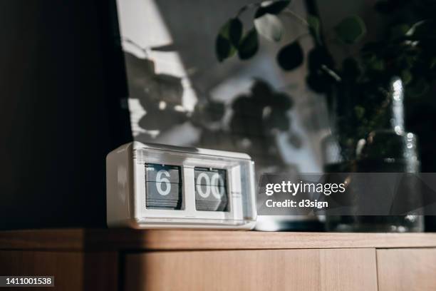 a white digital flip clock shows six o'clock on a brown wooden cabinet next to a fresh eucalyptus plant in the living room, with sunbeam shining through the window on a fresh beautiful morning. a brand new day, fresh start, fresh energy, new opportunities - alarm 個照片及圖片檔