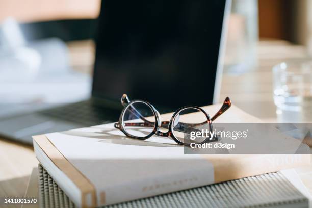 close up of eyeglasses on workspace table with books, documents and laptop in background with natural sunlight. office essentials. work from home. office workstation. freelance. self-employment concept - lunettes de vue photos et images de collection