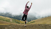 Hyped cheerful young woman jumping on grassy mountains surrounded by clouds