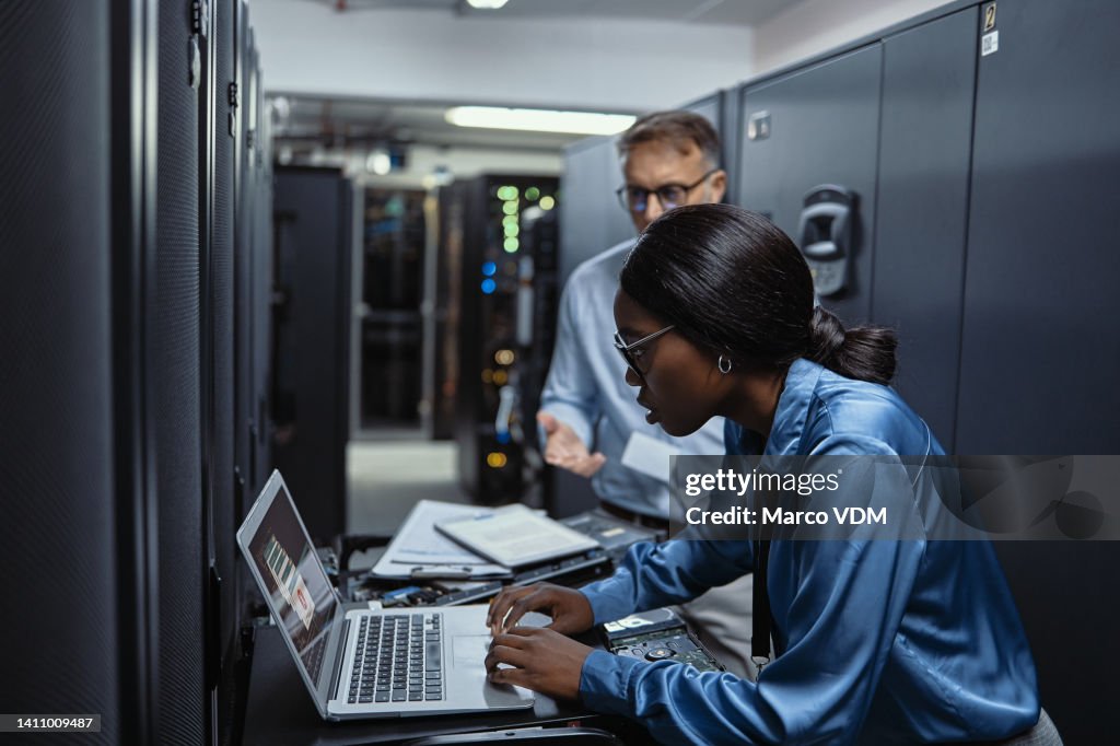 Two IT technicians using a laptop in a server room. Programmers fixing a computer system and network while doing maintenance in a datacenter. Engineers updating security software on a machine