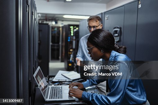 Two IT technicians using a laptop in a server room. Programmers fixing a computer system and network while doing maintenance in a datacenter. Engineers updating security software on a machine Two IT technicians using a laptop in a server room. Programmers fixing a computer system and network while doing maintenance in a datacenter. Engineers updating security software on a machine