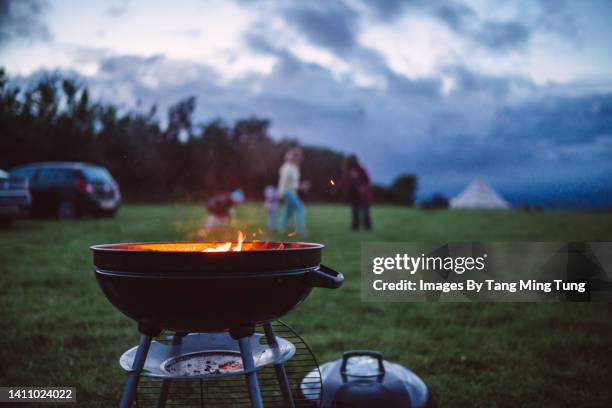 close up of a burning campfire at dusk in camping field with family having fun & playing in background - wood burning stove stock-fotos und bilder