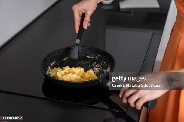unrecognizable female cooking scrambled eggs on frying pan on dark induction stove top. dark utensil in modern kitchen. faceless woman in casual orange dress preparing healthy food at home. horizontal, copy space, cropped - turner contemporary stock pictures, royalty-free photos & images
