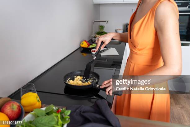 faceless woman cooking scrambled eggs on pan on induction stove top. dark utensil in modern scandinavia white kitchen. unrecognizable female in orange dress preparing healthy food at home. vegetables on plate. horizontal, copy space, cropped - stir frying european stock pictures, royalty-free photos & images