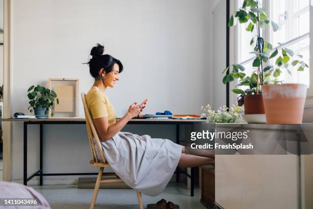 a smiling caucasian female businesswoman texting on her mobile phone while sitting at home - people mobilephone stockfoto's en -beelden