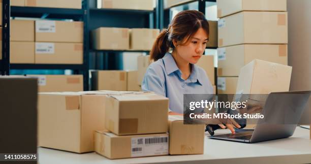 mujer de negocios asiática sentada frente al estante uso de código de barras escanear datos del cliente en caja de papel en el escritorio registro de código en línea detalle de pedido en computadora portátil para entrega urgente en el almacén. - material médico fotografías e imágenes de stock