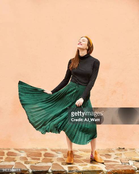 happy young woman standing outdoors in front of peach-coloured wall - long skirt stock pictures, royalty-free photos & images