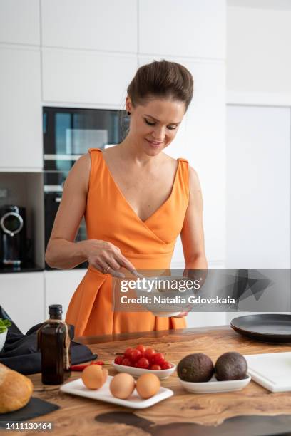 cheerful female stirring mixed eggs with spatula in bowl while cooking healthy nutritious morning breakfast in modern kitchen. preparing balanced omelet with tomatoes, avocados and olive oil. vertical - stir frying european stock pictures, royalty-free photos & images