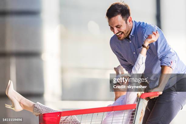 playful business couple having fun with shopping cart on the street. - man pushing cart fun play stock pictures, royalty-free photos & images