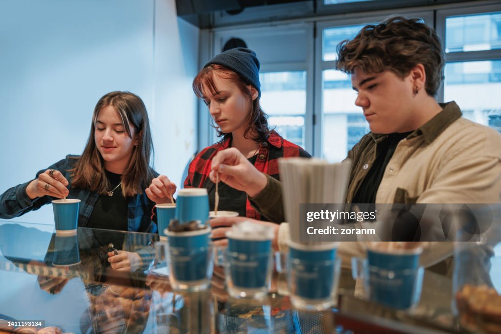 Three teenage friends stirring coffee to go in paper cups at the counter in cafeteria