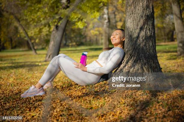 asian woman sitting on ground leaning on a tree, smiling towards sunshine, holding water bottle - leaning tree stock pictures, royalty-free photos & images