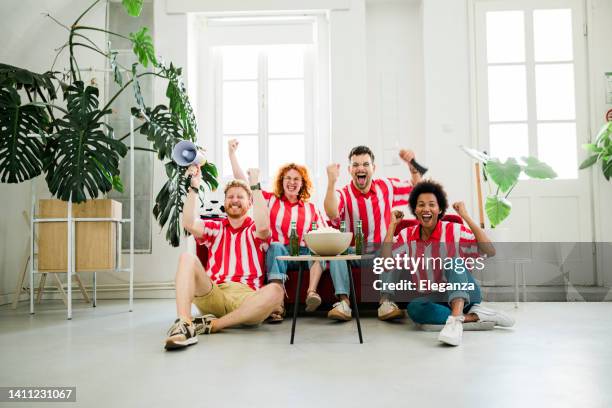 excited friends cheering while watching football match on sofa at home - basketball match on tv stockfoto's en -beelden