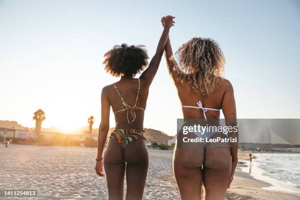 two young generation z women holding hands on the beach looking at susnet - couple swimwear stock pictures, royalty-free photos & images