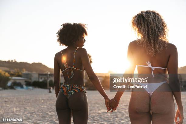 two young generation z women holding hands on the beach looking at susnet - black girl swimsuit stock pictures, royalty-free photos & images