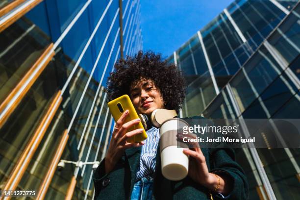low angle view of woman standing between commercial buildings using mobile phone. - groovy fotografías e imágenes de stock