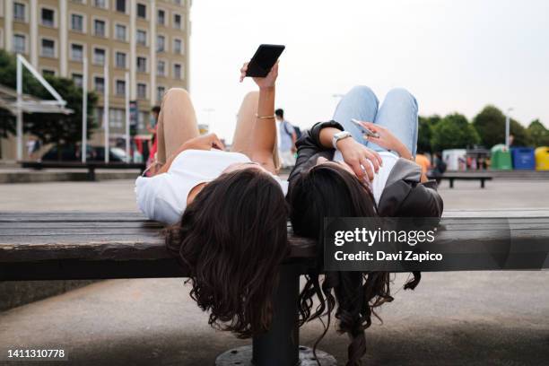 two unrecognizable women laying down on a bench in the street and looking at phone. - girl lying down stock pictures, royalty-free photos & images