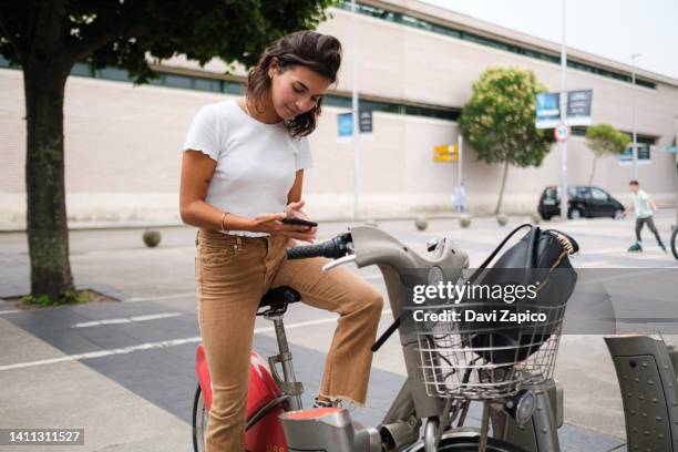 young latin woman looking at phone on the bike rental parking. - bicycle rental stock pictures, royalty-free photos & images