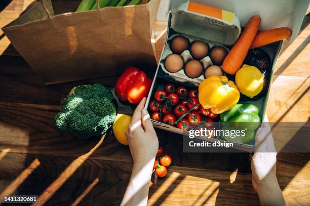 cropped hands of woman opening a home delivery cardboard box filled with multi-coloured fresh organic fruits, vegetables and groceries on wooden background against sunlight. grocery subscription boxes. meal kits. convenient and time savings for daily life - plastic free stock pictures, royalty-free photos & images