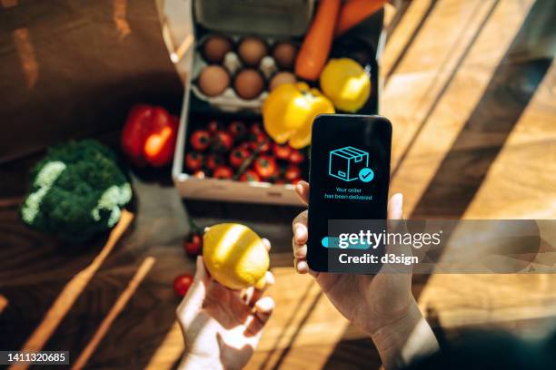 female hands confirming the arrival of a box of home delivery filled with multi-coloured fresh and healthy organic groceries ordered online  with mobile app on smartphone. grocery subscription boxes. meal kits. convenient and time savings for daily life - asien e commerce stock-fotos und bilder