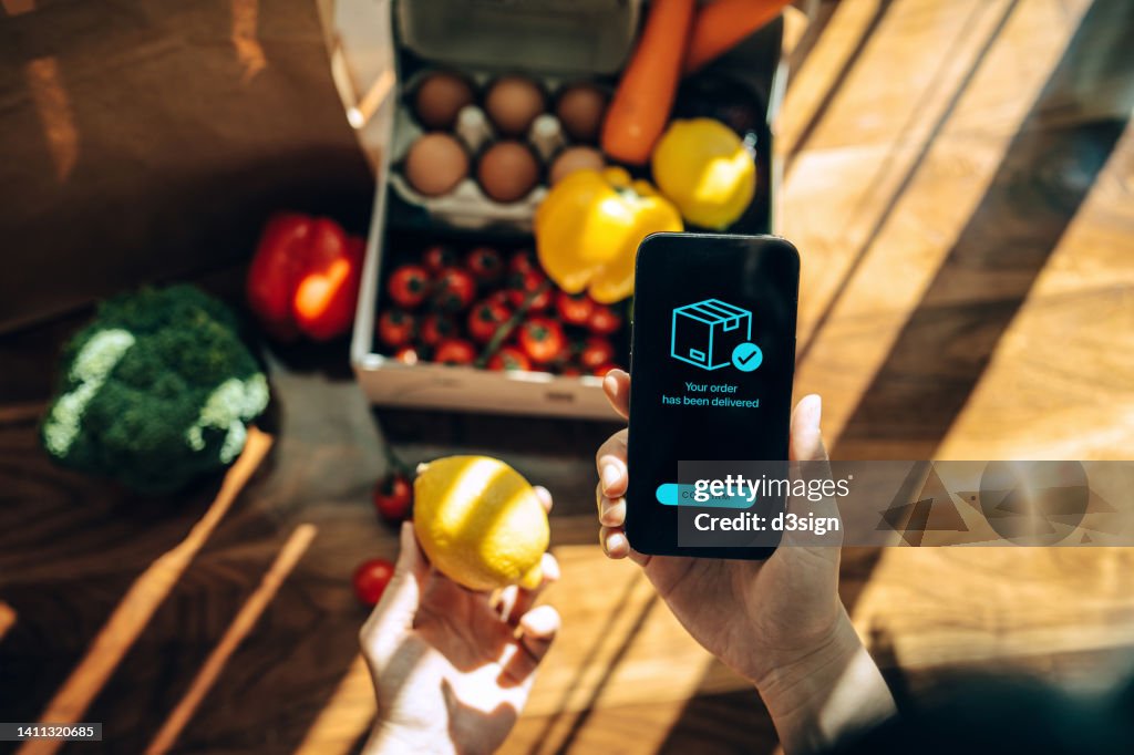 Female hands confirming the arrival of a box of home delivery filled with multi-coloured fresh and healthy organic groceries ordered online with mobile app on smartphone. Grocery subscription boxes. Meal kits. Convenient and time savings for daily life