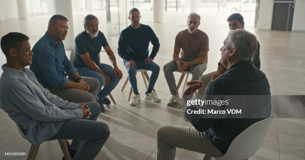 A team of men sitting in a circle during a therapy session. Diverse colleagues talking about their emotions in a support group with a professional psychologist in a office or mental health workshop