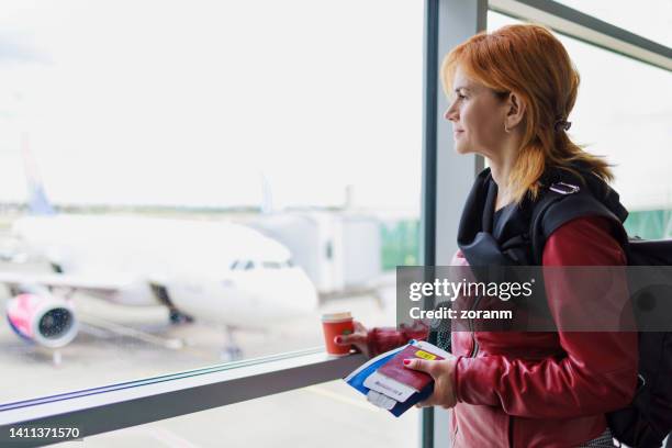 woman standing by the window in airport departure area, having coffee in paper cup and looking outside - airplane tickets stock pictures, royalty-free photos & images