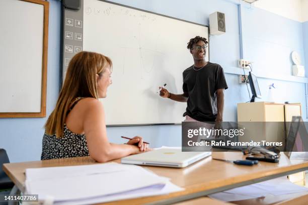 black teen male student explaining exercise on chalkboard  to classroom mates - two female teachers blackboard stockfoto's en -beelden