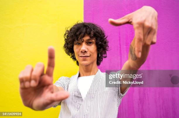 jovem gay sorrindo através de uma moldura de dedo contra um fundo colorido - dedos fazendo moldura - fotografias e filmes do acervo