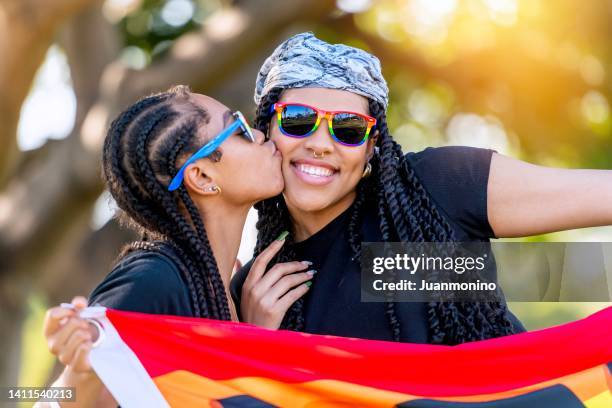 afro-latinx young lesbian kissing her wife holding a rainbow banner with the word proud written on it - black lesbians kiss stock pictures, royalty-free photos & images