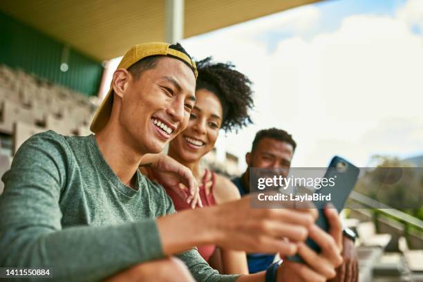 people outdoors smiling at phone, dressed casually. young, diverse group looking and laughing at website on mobile excitedly. friends sitting with each other on stadium chairs - diverse group of people outside stock pictures, royalty-free photos & images