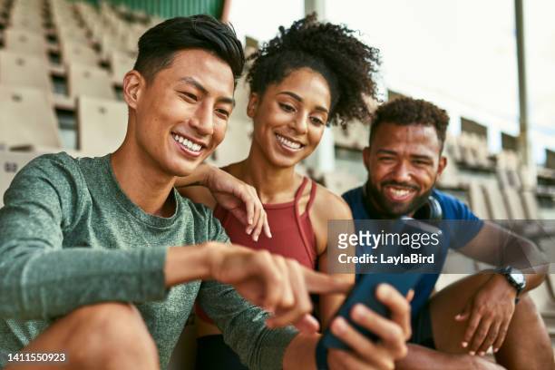 group of athletes tracking fitness using a sports app on a phone, logging their cardio stats online while sitting together in a stadium. sporty friends laughing during a video call online - liberty stadion stock pictures, royalty-free photos & images