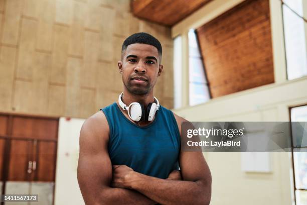 fit and healthy athlete ready for a workout session in indoor gym court with headphones around his neck. portrait of black male serious about fitness. handsome sports coach standing with folded arms - headphone man on neck stock pictures, royalty-free photos & images