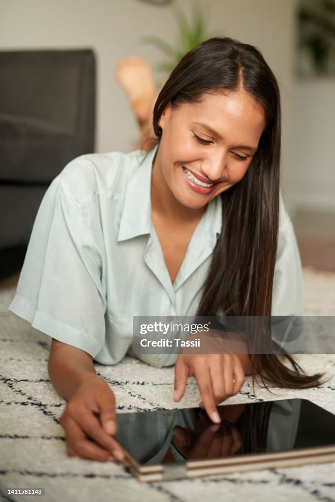 A young woman relaxing and watching videos on a tablet while lying on the floor at home. Female watching video on a digital device resting in her house on a weekend. Lady enjoying some entertainment