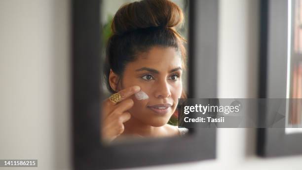 woman applying moisturizer to her face while grooming in the mirror at home. young female using sunscreen with spf for uv protection. wearing lotion daily for healthy complexion and glowing skin - perfect complexion stock pictures, royalty-free photos & images