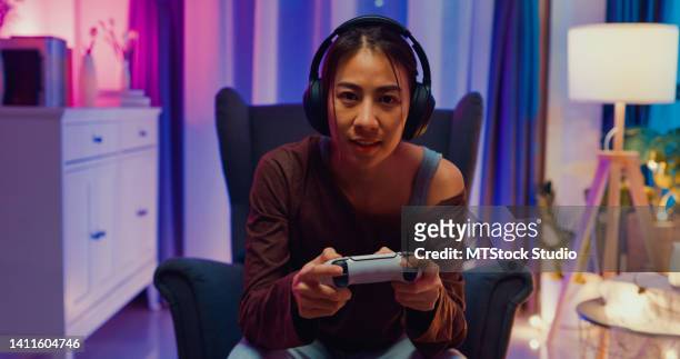 happy young asian woman wear casual sit on sofa chair hold joystick controller exciting and shouting while having fun winning playing video game sit on sofa in the living room home at night. - game streamer stock pictures, royalty-free photos & images